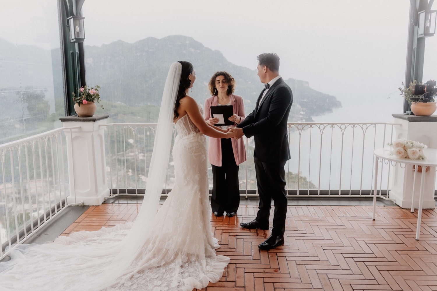 A couple exchanging vows with their celebrant on a terrace at Palazzo Avino in Ravello, with the Amalfi Coast behind them