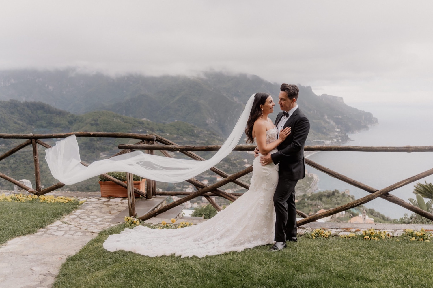 A couple embracing on a grassy terrace above the Amalfi Coast at Palazzo Avino in Ravello, with the bride's long veil in the wind and the coast behind them