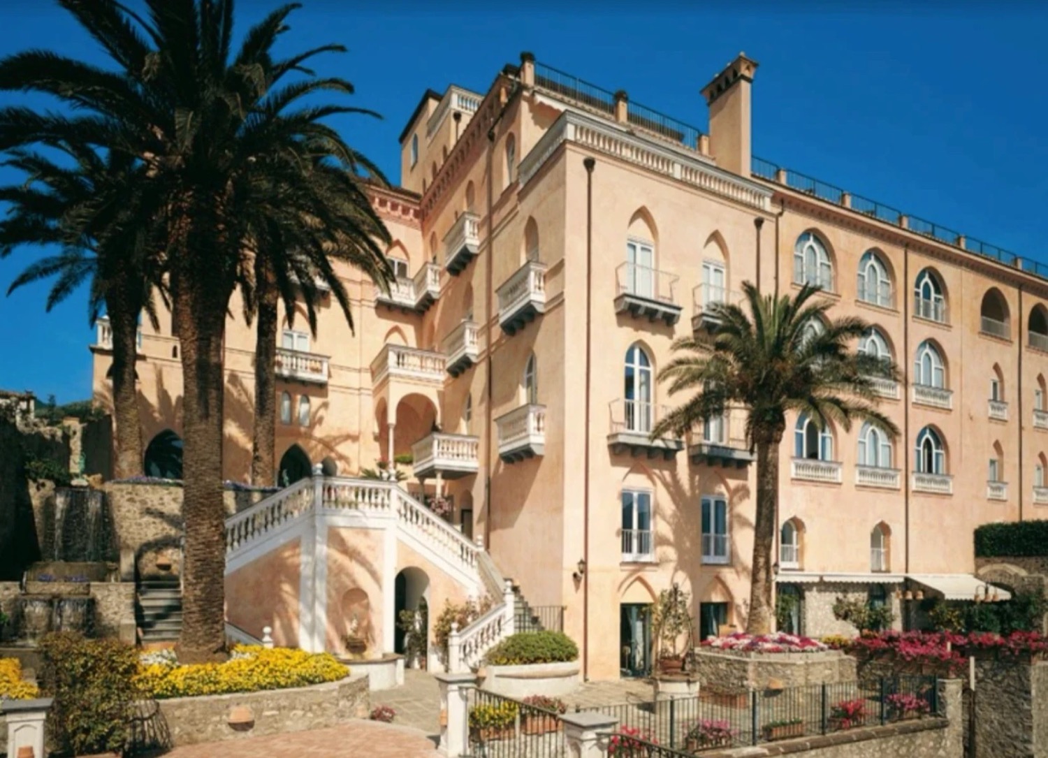 The pink facade of Palazzo Avino in Ravello, with palm trees, a white balustraded staircase and flower beds