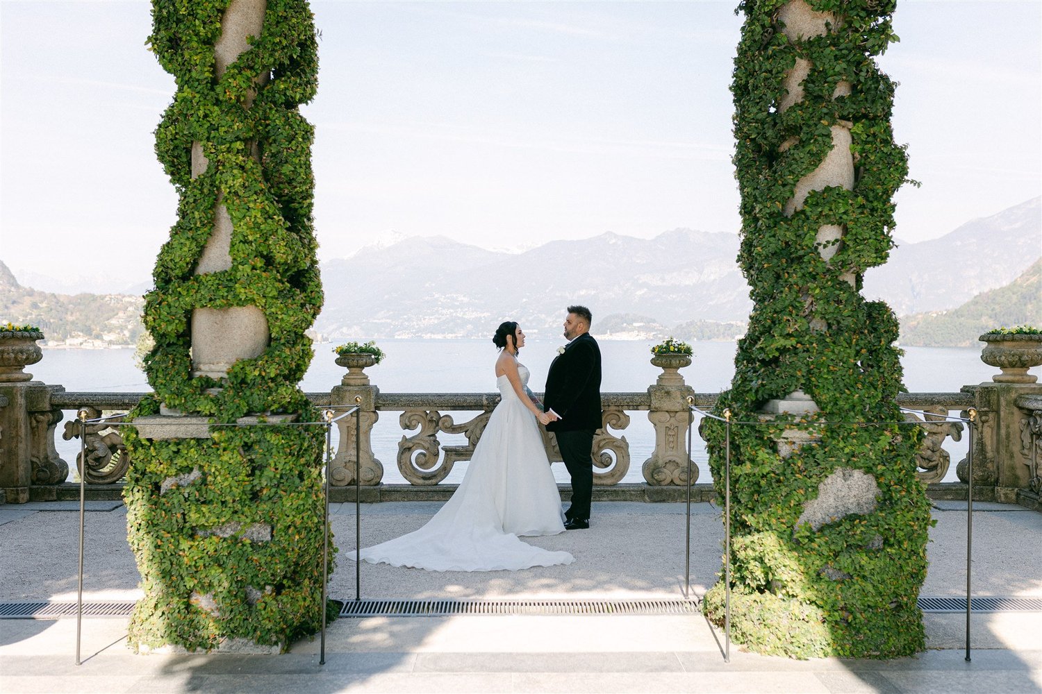 A couple holding hands between the green-clad arches of the Loggia Durini at Villa del Balbianello, with Lake Como behind