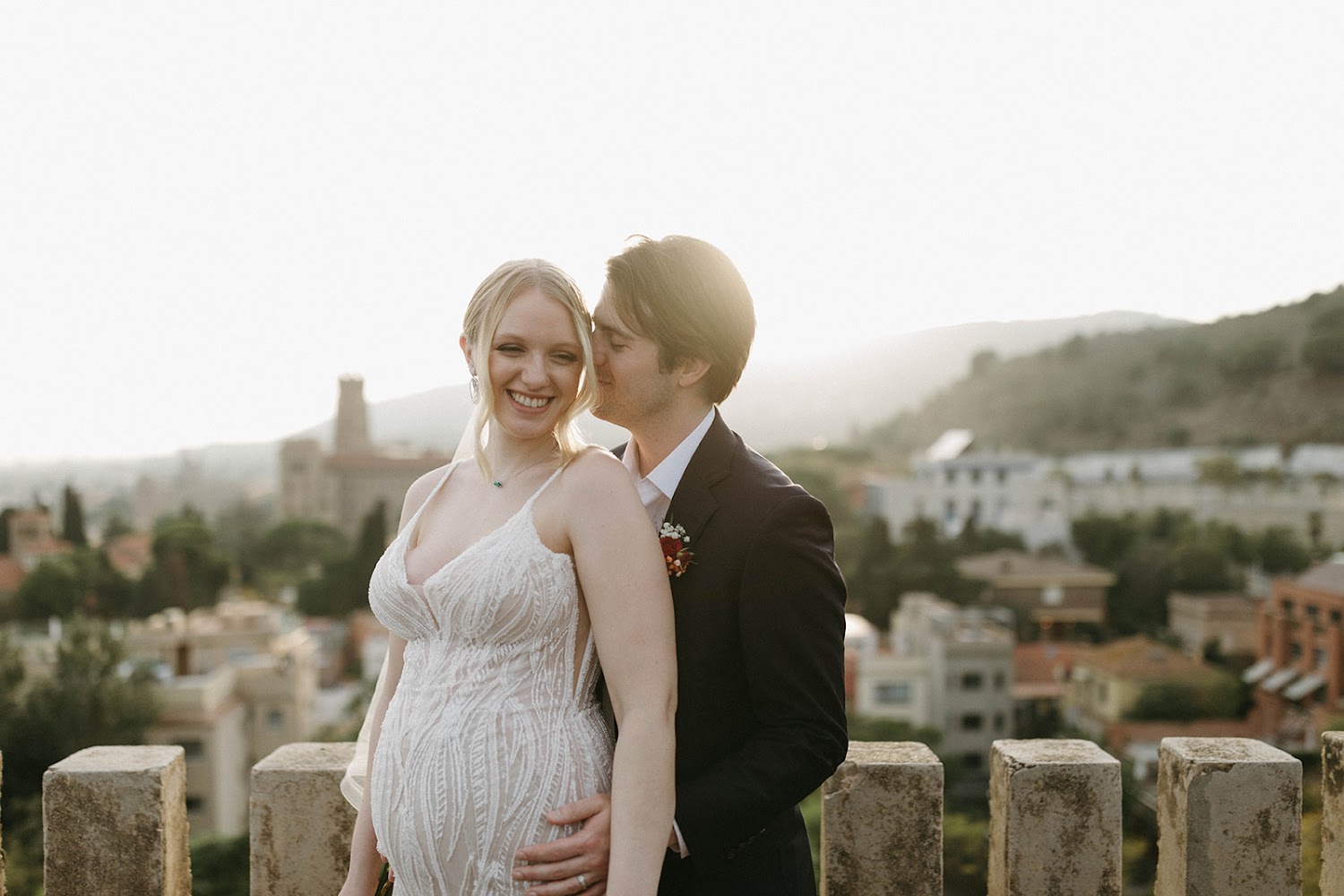 A smiling couple embracing on the stone terrace of Bellesguard Tower, a Gaudí building in Barcelona, with the city and hills of Barcelona behind them