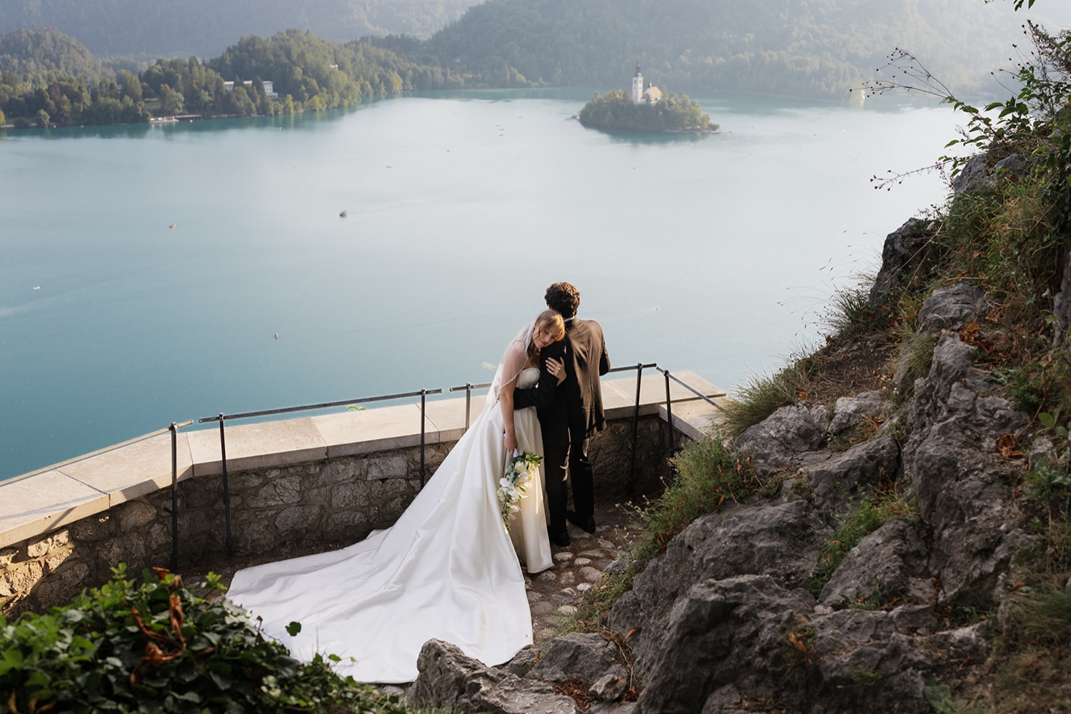 A bride resting her head on her groom's shoulder on the terrace of Bled Castle, with Lake Bled and its island below, Slovenia