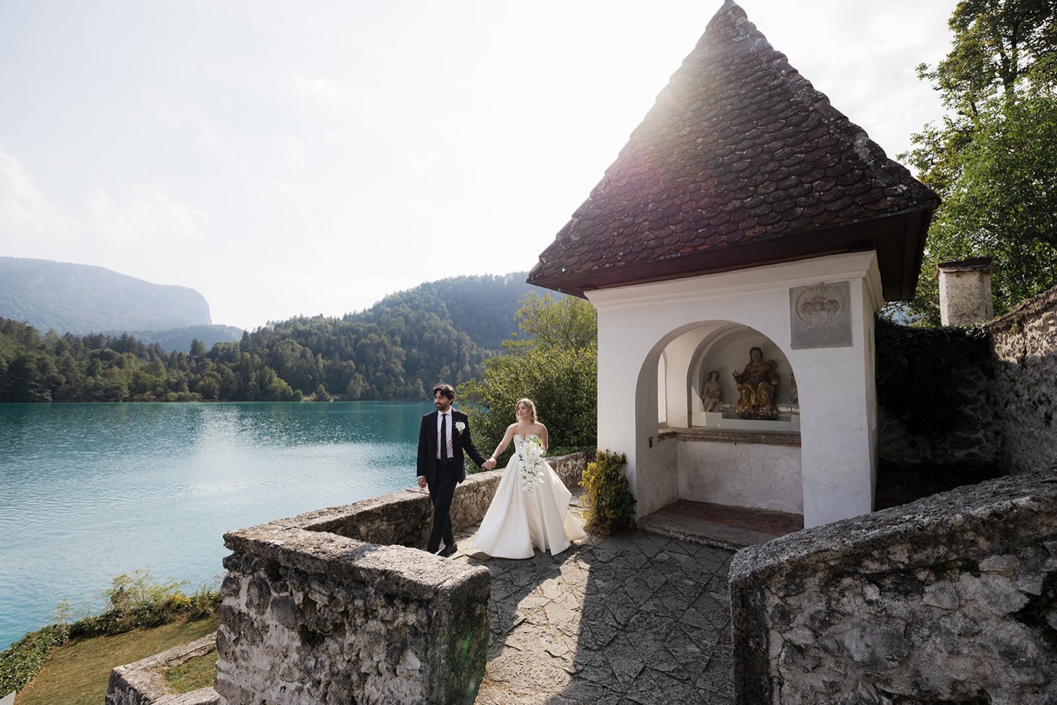 A couple walking hand in hand beside a small chapel at Bled Castle, with Lake Bled beside them, Slovenia