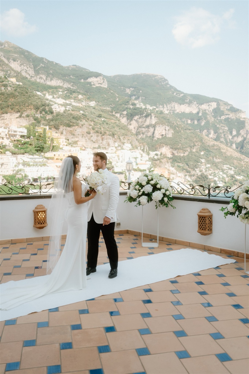 A bride and groom at the end of the white aisle on the rooftop terrace of Hotel Conca d’Oro in Positano, with the mountains behind them