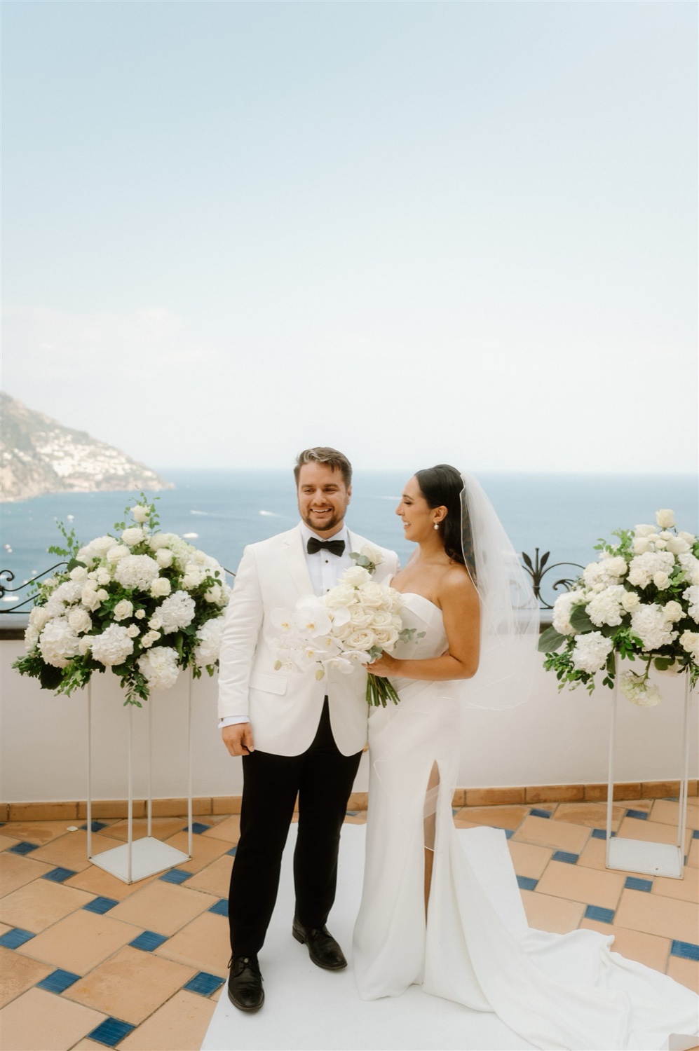 A bride and groom between white flower arrangements on the rooftop terrace of Hotel Conca d’Oro in Positano