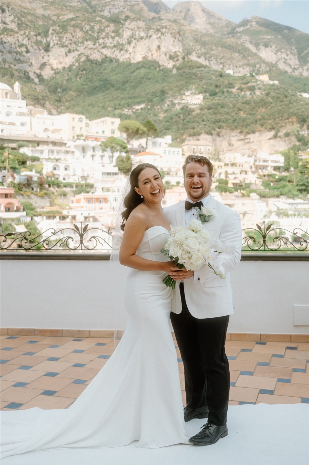 A couple laughing together on the rooftop terrace of Hotel Conca d’Oro in Positano, with the village and mountains behind