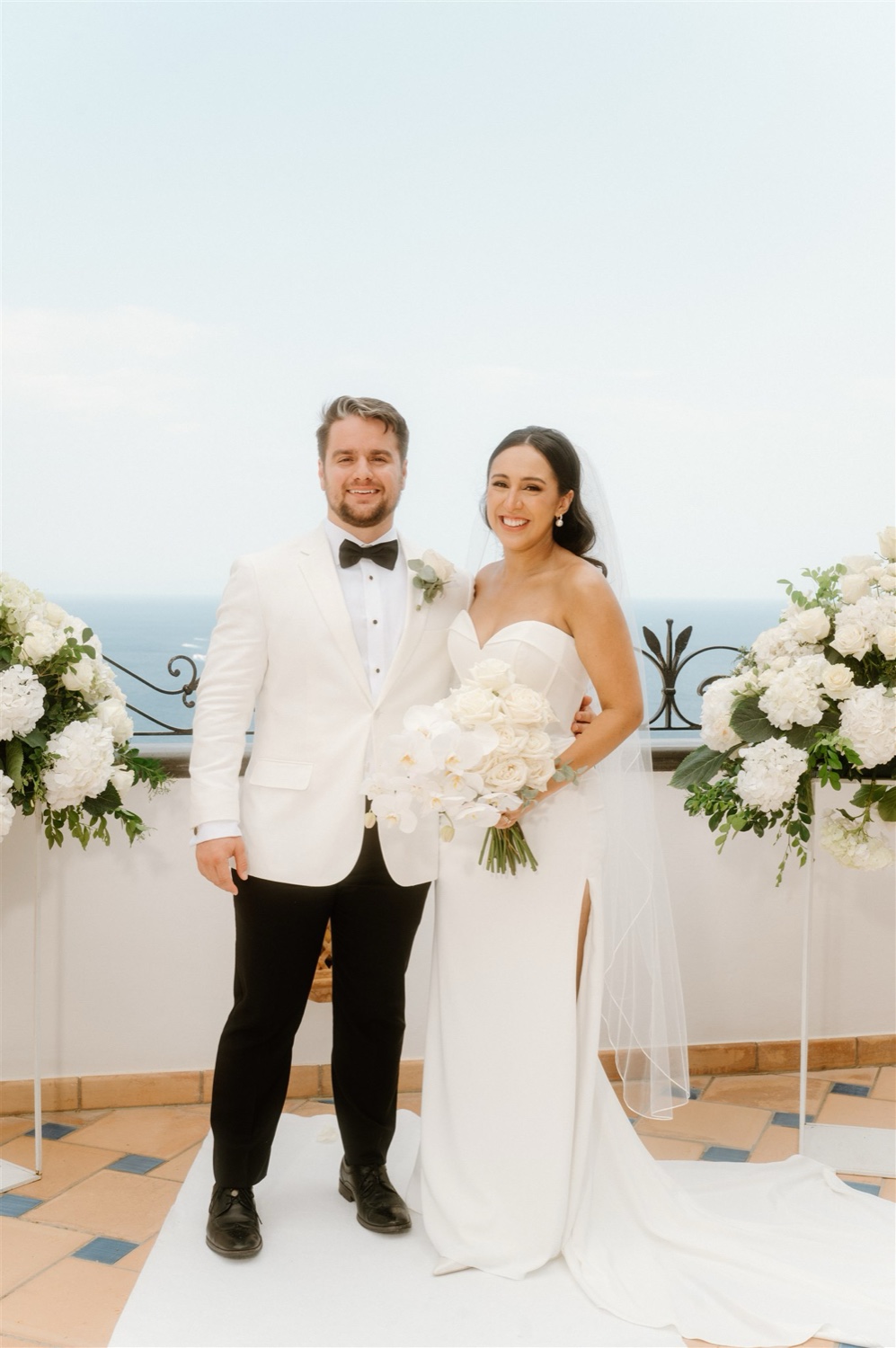 A bride and groom standing together on the rooftop terrace of Hotel Conca d’Oro in Positano, with white flowers and the sea behind