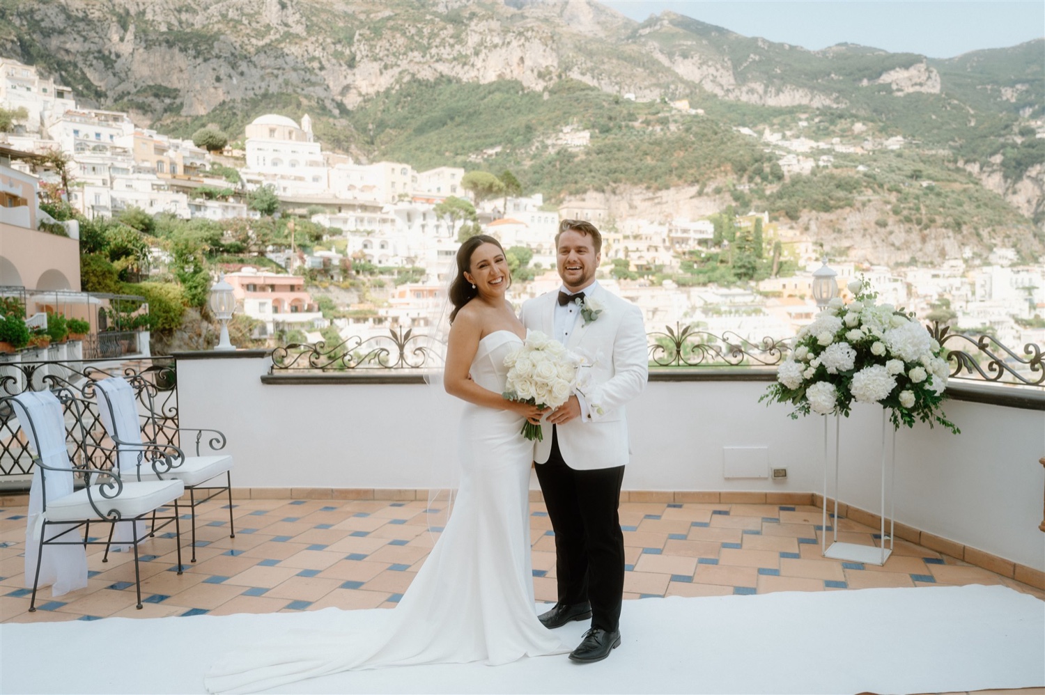 A bride and groom smiling on the rooftop terrace of Hotel Conca d’Oro in Positano, with the village and the mountains behind them
