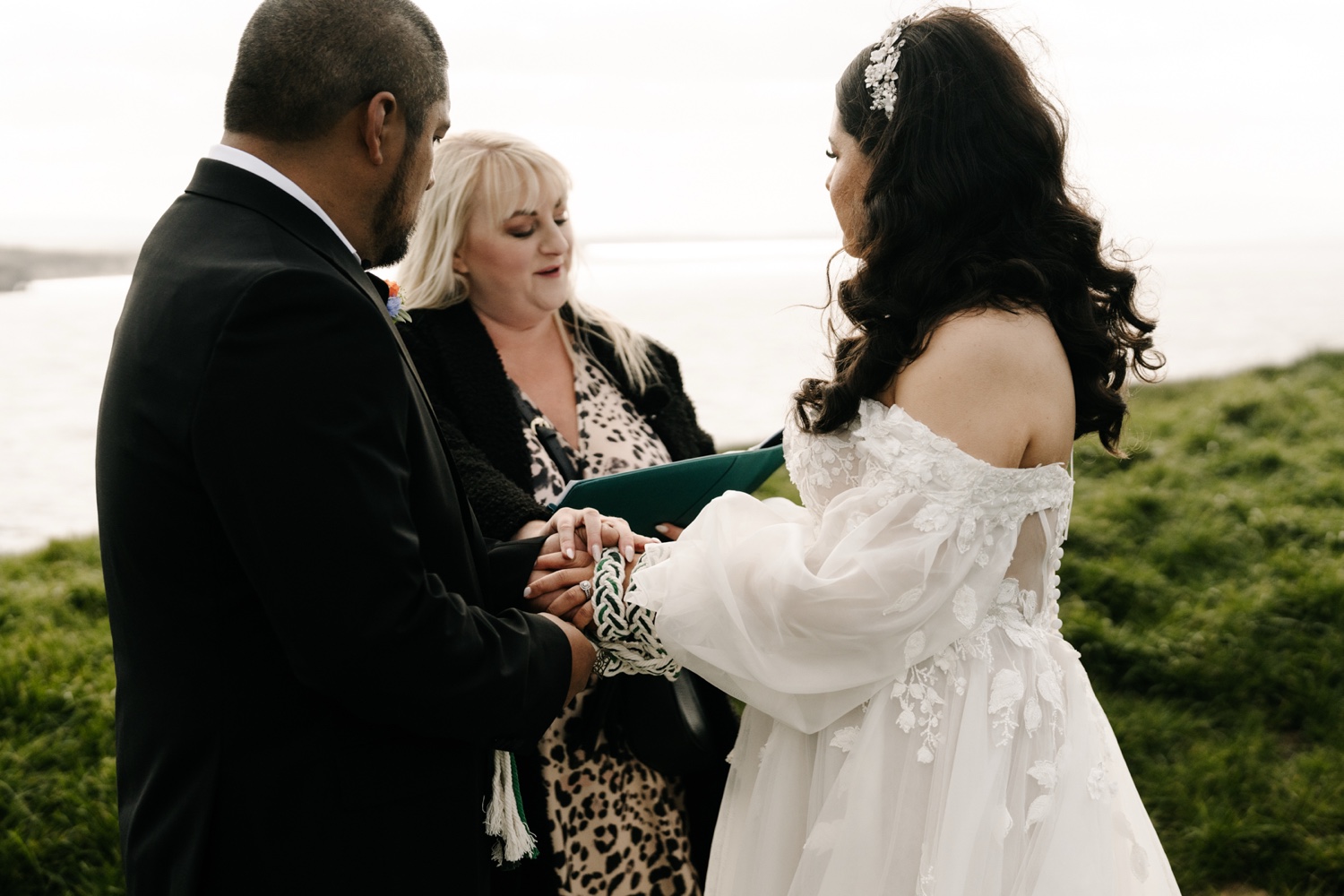 A couple exchanging rings with their celebrant in a clifftop field near Dunluce Castle, Northern Ireland