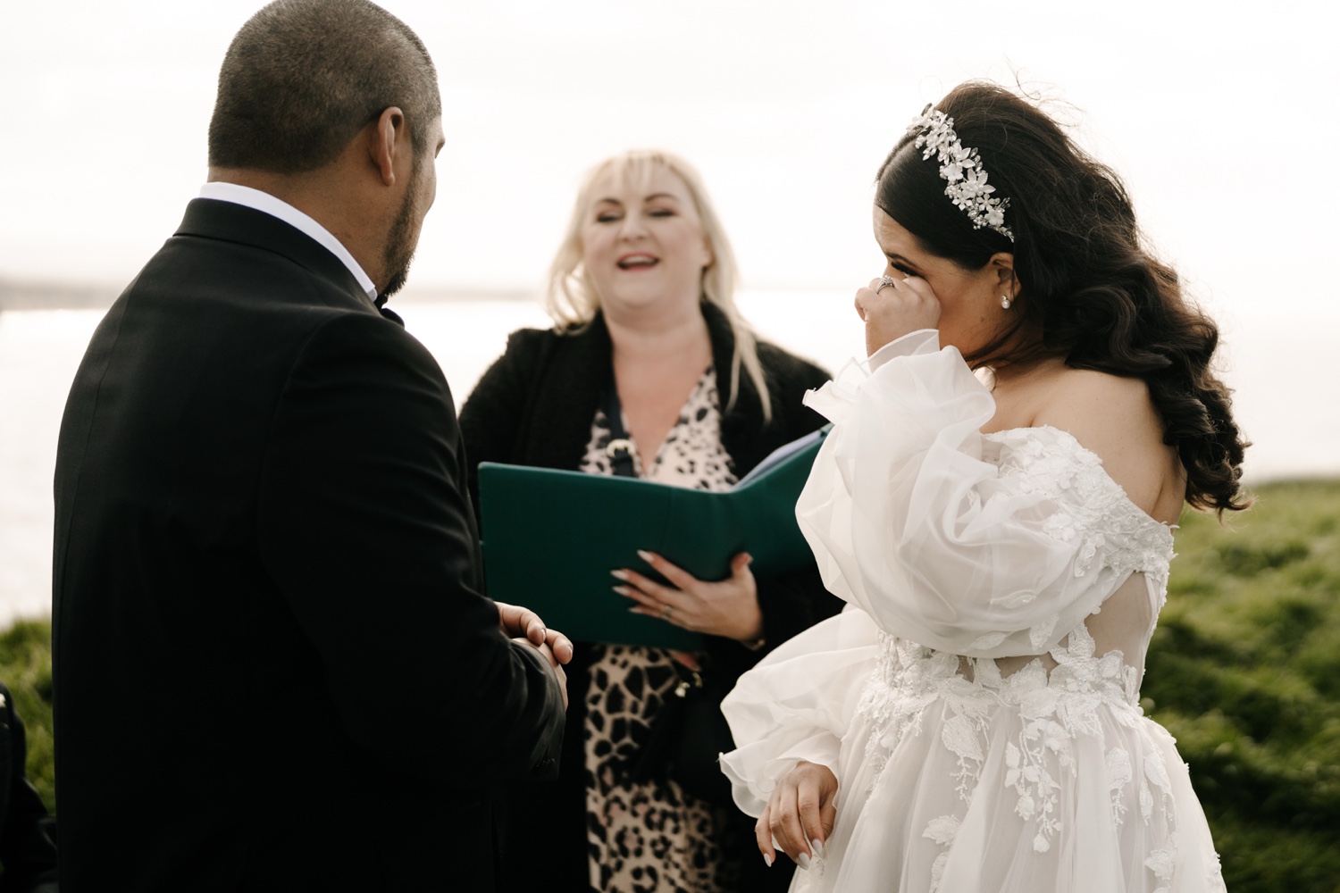 A bride wiping away a tear as she exchanges rings with her groom and celebrant near Dunluce Castle, Northern Ireland
