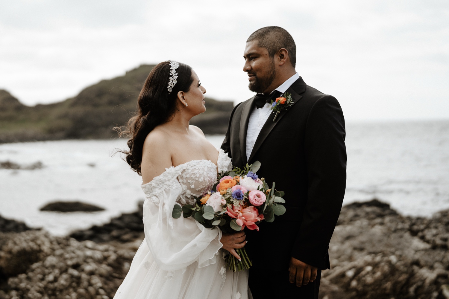 A bride with a colourful bouquet and her groom looking at each other by the sea on the north coast of Northern Ireland