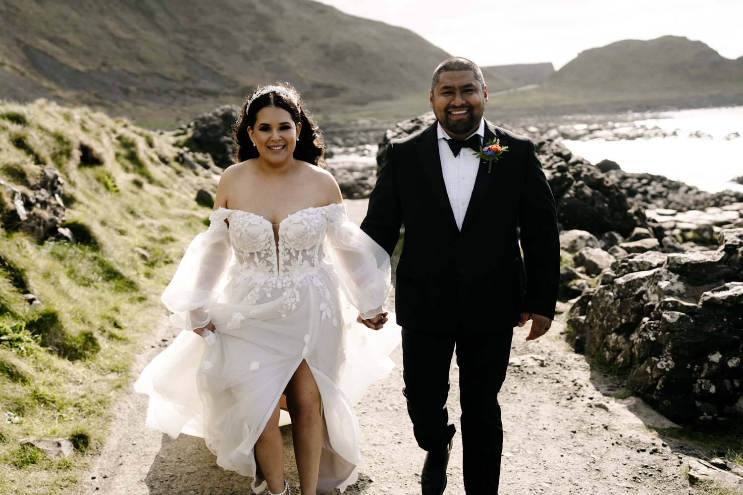 A smiling bride and groom walking hand in hand along a path above the sea in Northern Ireland