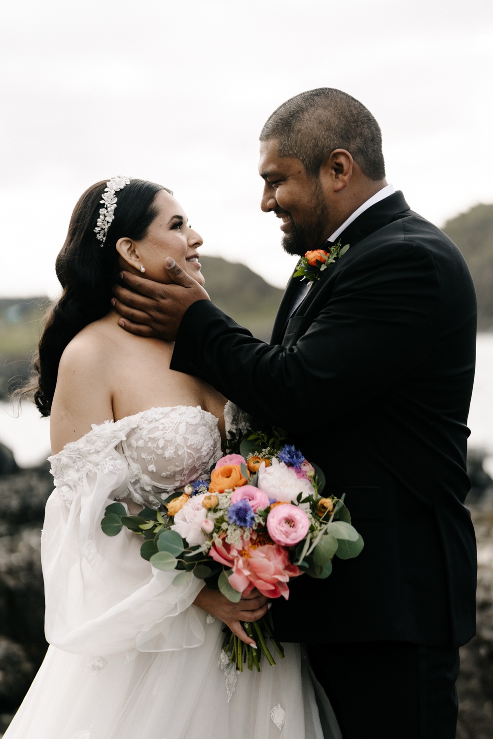 A groom holding his bride's face as they look at each other with a bouquet by the sea in Northern Ireland