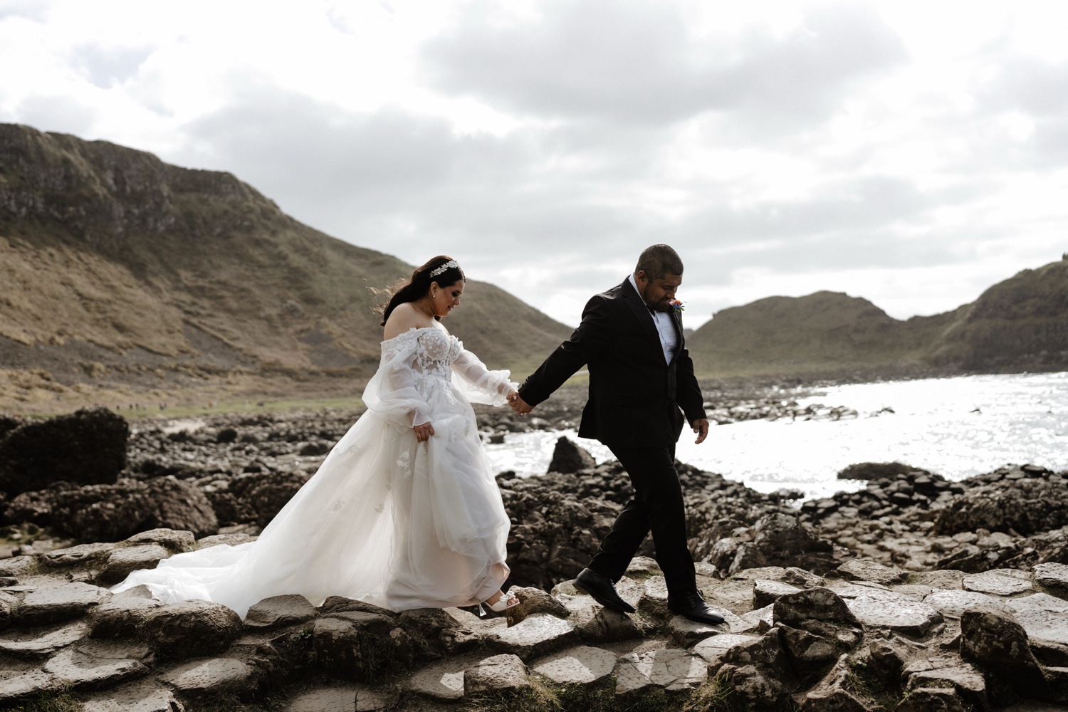 A groom helping his bride across the rocks beside the sea in Northern Ireland