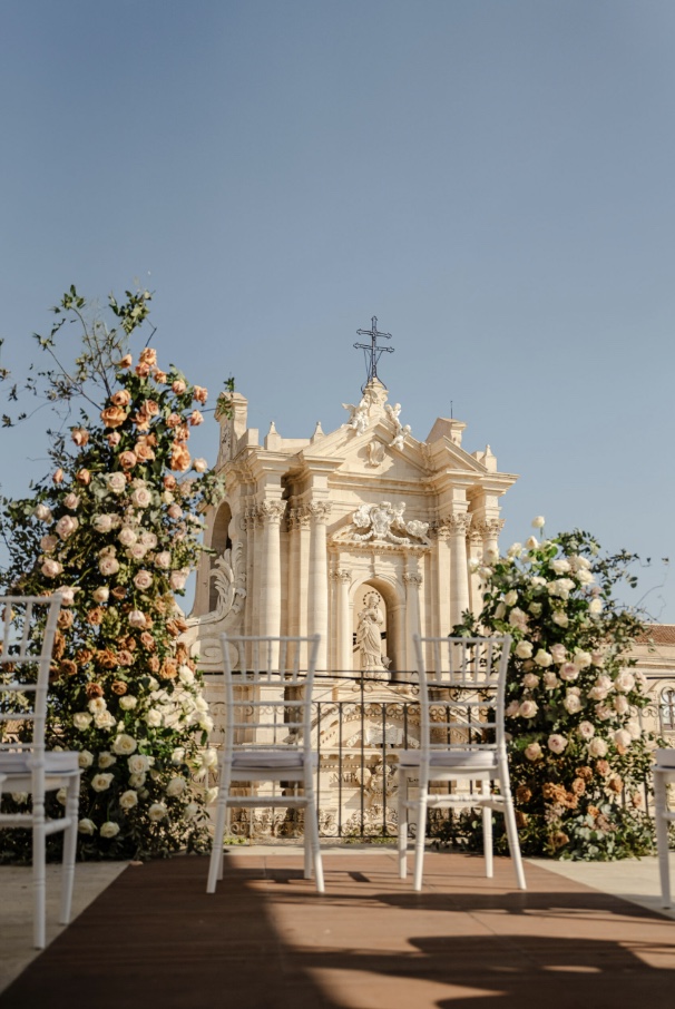Elopement ceremony setup with white chairs and peach and cream rose arrangements on the Terraza Killiche terrace, facing the baroque church in Sicily