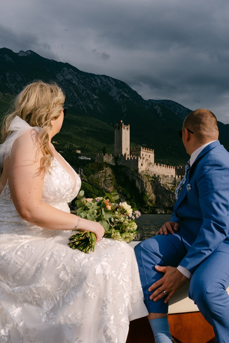 A bride and groom on a boat with the castle of Malcesine on the cliff behind them