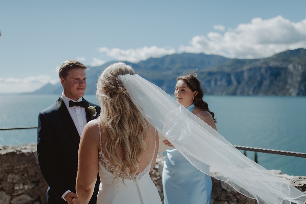 A groom smiling at his bride as her veil blows in the wind during their vows on a castle terrace in Malcesine above Lake Garda