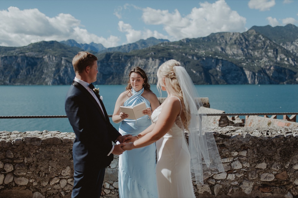A bride and groom holding hands as their celebrant reads during a symbolic elopement ceremony on the Scaliger Castle terrace in Malcesine, Lake Garda
