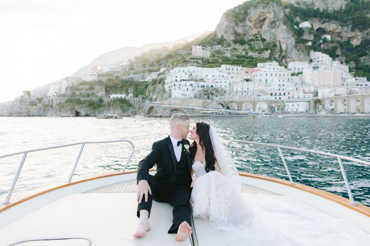 Newlyweds sitting on the bow of a private boat in front of Amalfi's white houses