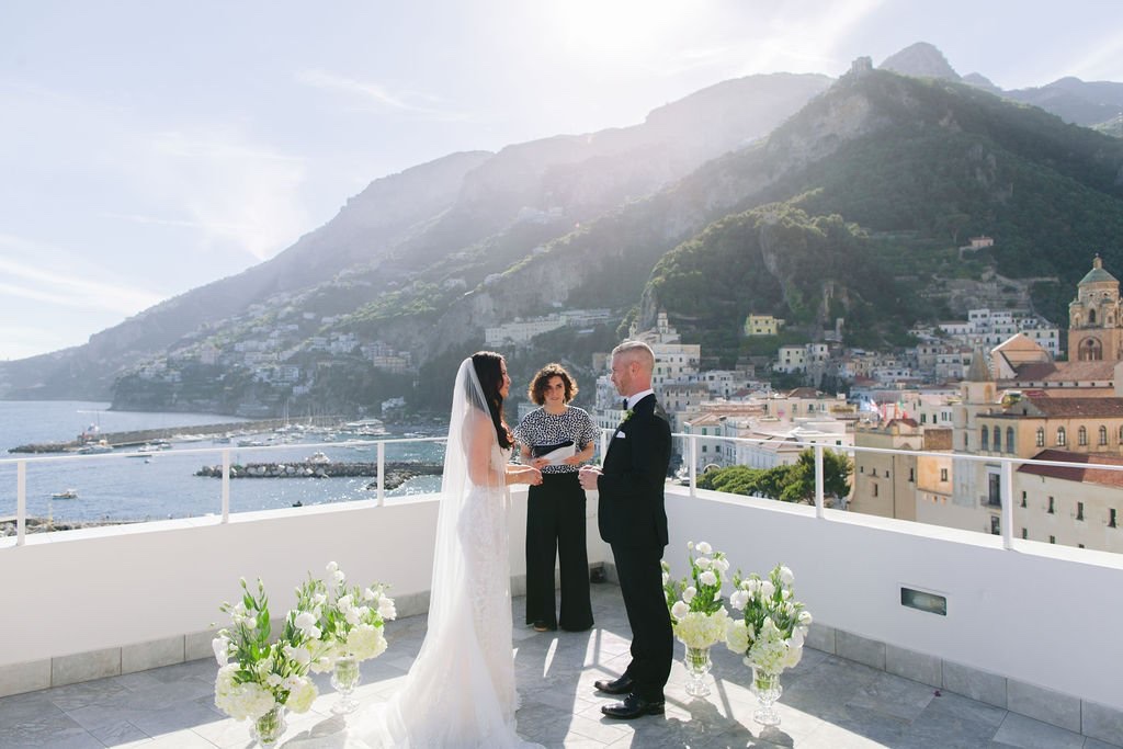 A couple exchanging vows with their celebrant on the white rooftop terrace of Hotel Marina Riviera, with the Amalfi Coast mountains and the town of Amalfi behind