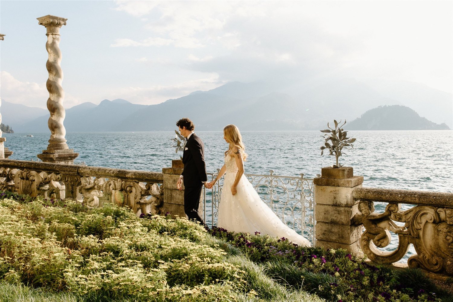 A couple walking along the balustrade of the gardens of Villa Monastero, with Lake Como and the mountains behind
