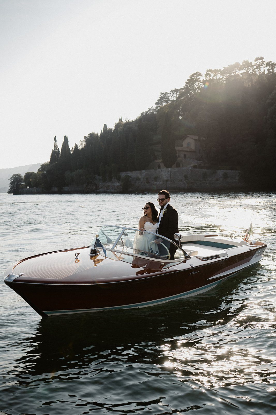 A couple crossing Lake Como on a wooden boat towards a wooded shore