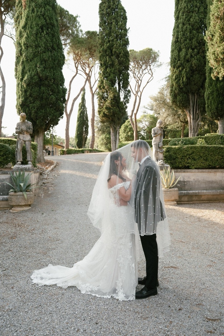 A bride and groom under a long veil on the gravel garden path at Villa Poggiano, Tuscany