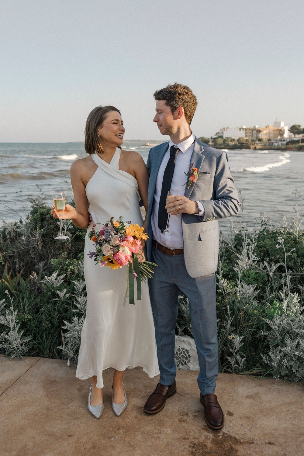 A bride and groom smiling together with a bouquet and a glass of champagne by the sea at Restaurant Sa Punta in Mallorca