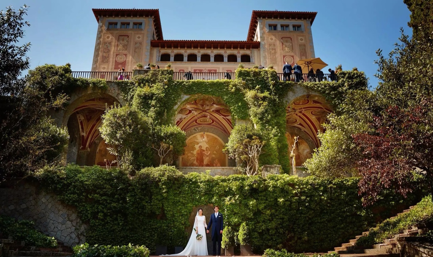 A couple standing at the foot of the ivy-covered terraces of Bell Reco, a historic masia on the Costa Brava, with the stone house above them