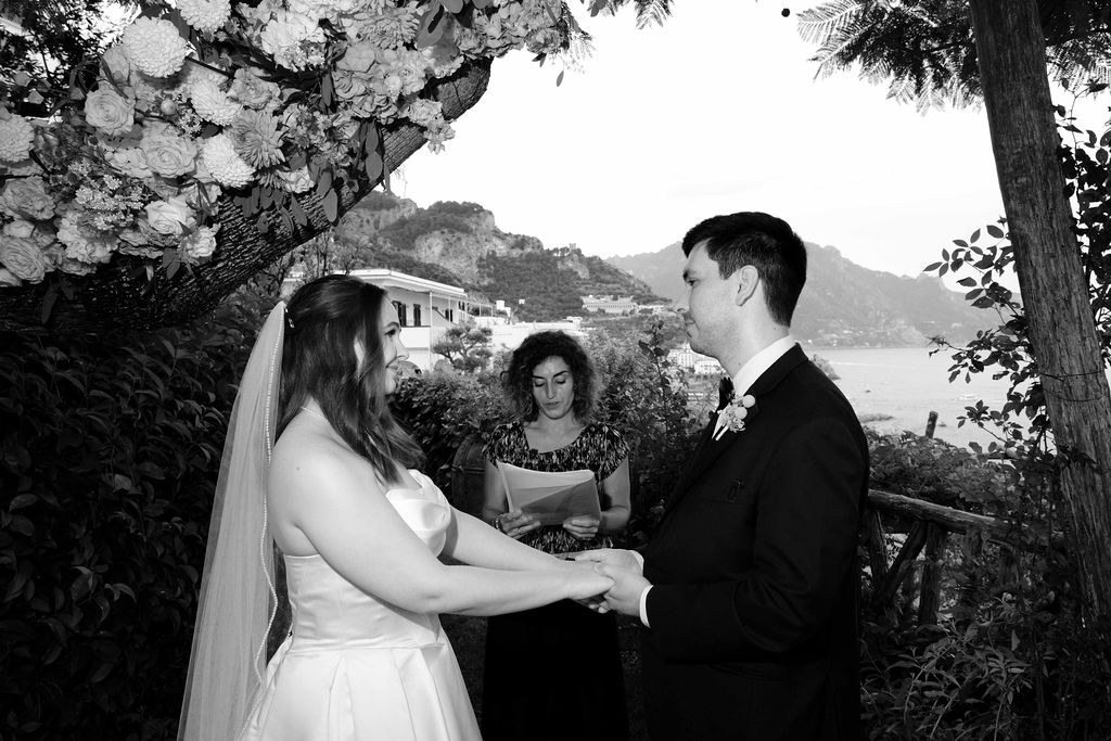 A couple holding hands during their vows in the garden of Hotel Santa Caterina, with the Amalfi Coast behind them, in black and white