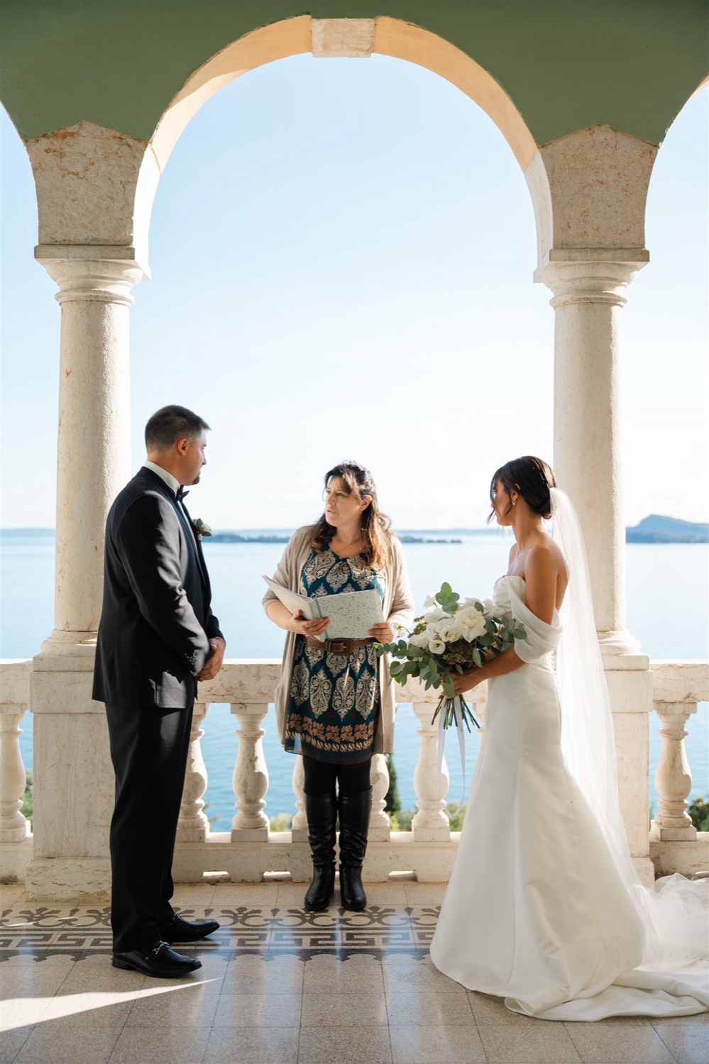 A bride and groom exchanging vows with their celebrant between stone columns on the terrace of Villa del Sogno on Lake Garda