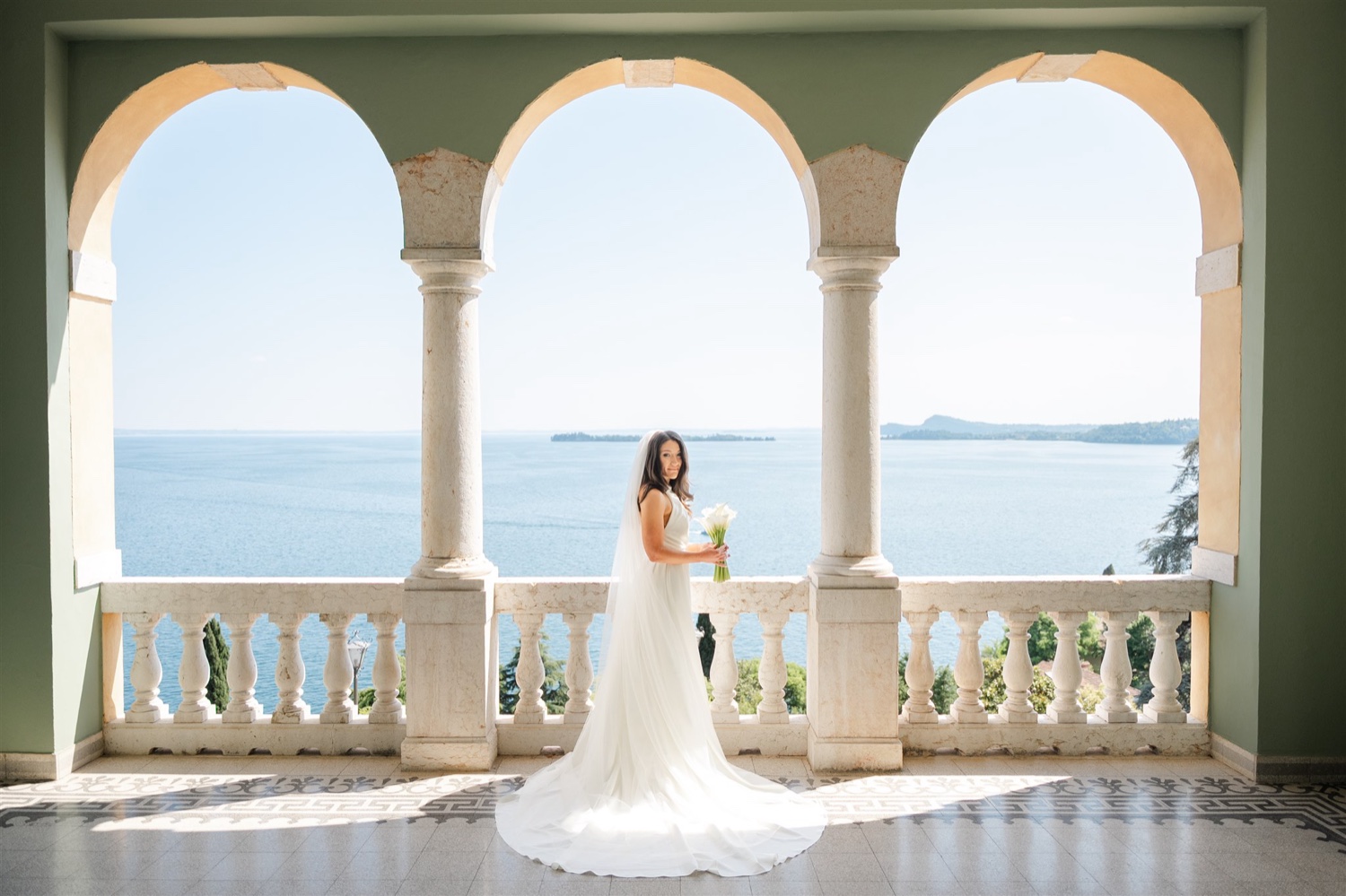 A bride standing on the arched terrace of Villa del Sogno on Lake Garda, with the lake and the far shore behind her