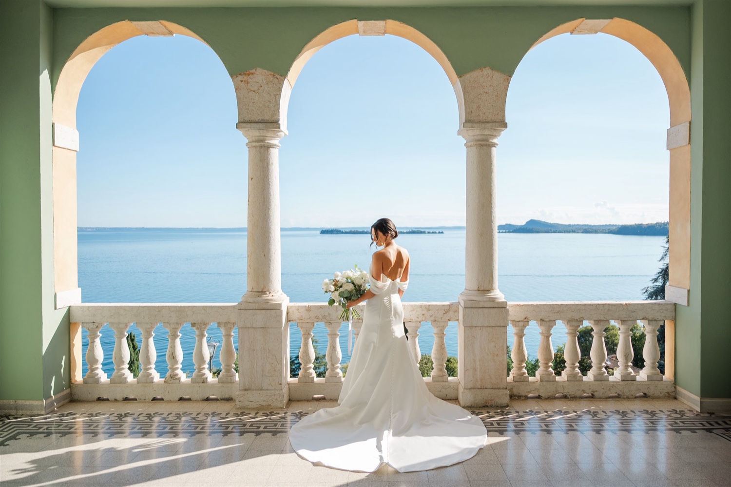A bride standing on the arched terrace of Villa del Sogno on Lake Garda, with the lake and a small island behind her