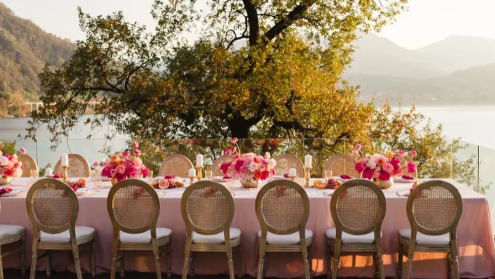 A long pink dining table with pink and orange flowers under a tree on the terrace at Villa Porta, Lake Maggiore, Italy