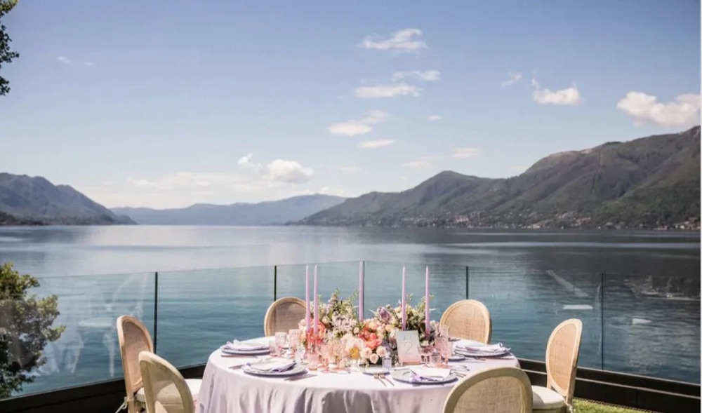 A round table set with lilac candles and flowers on the glass-fronted terrace at Villa Porta overlooking Lake Maggiore