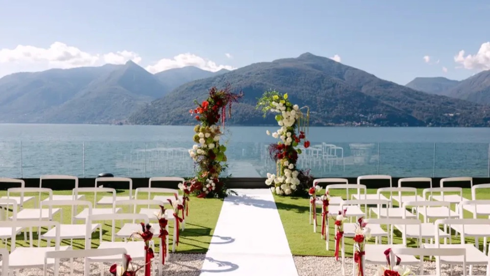A ceremony set up on the Villa Porta terrace with a red and white floral arch and a white aisle, mountains behind, Lake Maggiore, Italy