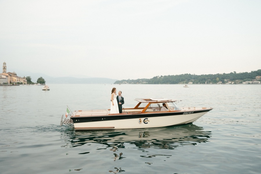 A bride and groom standing on a classic wooden boat on Lake Garda during their elopement in Italy