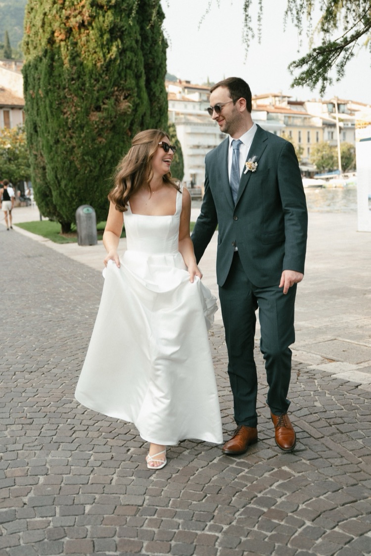 A bride and groom in sunglasses laughing as they walk along a lakeside promenade on Lake Garda, Italy