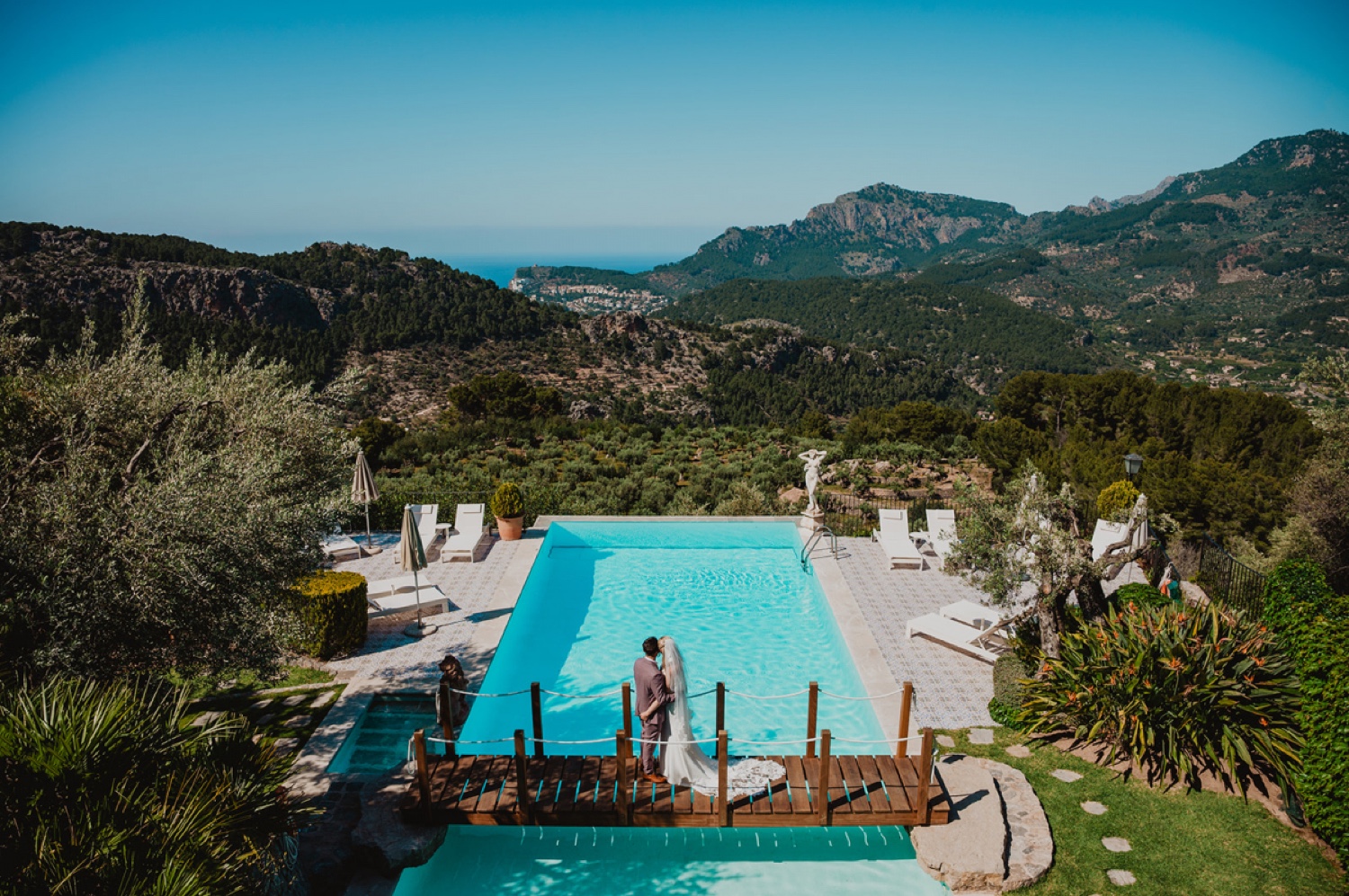 A couple on the wooden jetty of the turquoise pool at Ca’s Xorc, with the mountains and the sea of Mallorca beyond