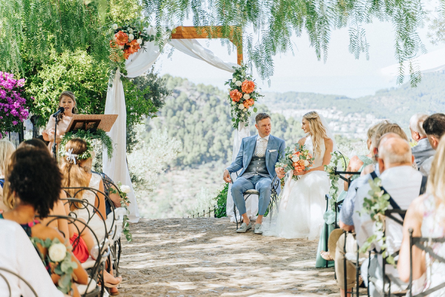 A couple seated under a floral arch for their ceremony in the gardens of Ca’s Xorc in Mallorca, with the mountains behind